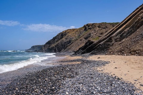 Waves hit a pebble beach, high rocky cliffs rise in the background under a clear sky, Rogil, Faro,