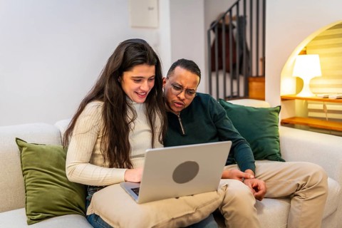 Happy young multiracial couple sitting on a cozy sofa at home, browsing content on a laptop screen,