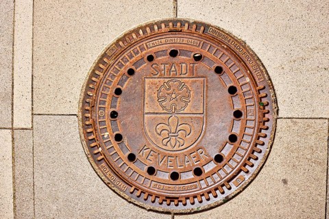Manhole cover city of Kevelaer, city coat of arms, footpath made of concrete paving stones, sunny,