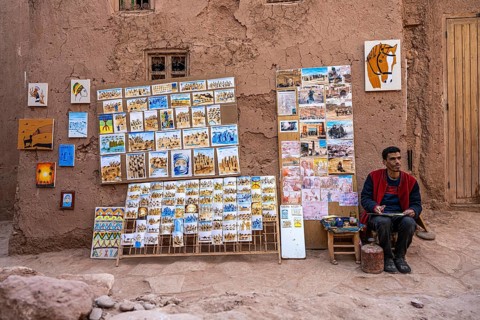 A Moroccan artist, painter exhibits his paintings for sale in front of a clay house, Kasbah Ait