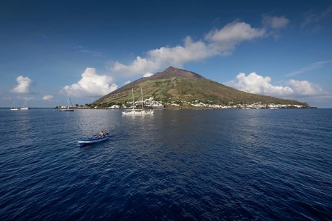 Island with volcano, boats on calm sea under blue sky, Stromboli, Aeolian or Lipari Islands,