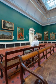 A visitor sits in a gallery in front of paintings surrounded by green walls and wooden benches,