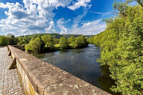 View of a river from a bridge, surrounded by green forest and under a blue sky with clouds, view of