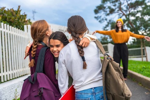 Young girls carrying backpacks and school supplies, smiling and embracing casually on the sidewalk,
