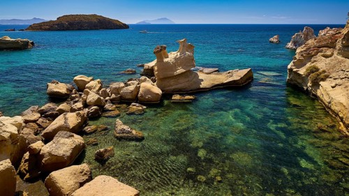 A variety of rocks rise from crystal clear water against a small island in the background,