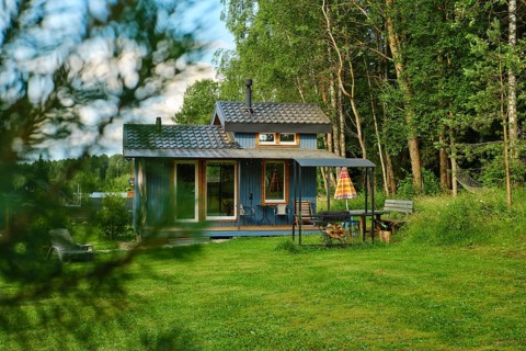 Wooden house stands in green field surrounded by tall trees. Table, grill and chair are set outside