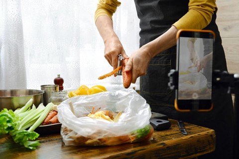A person is peeling carrots while standing at a wooden table in a kitchen.He records a video on his