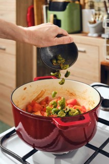 Hand is adding crushed green olives to pot filled with chopped tomatoes and vegetables. Pot sits on