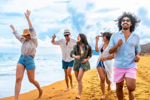 Diverse young friends running barefoot along a sunny sandy beach by the ocean, smiling and laughing
