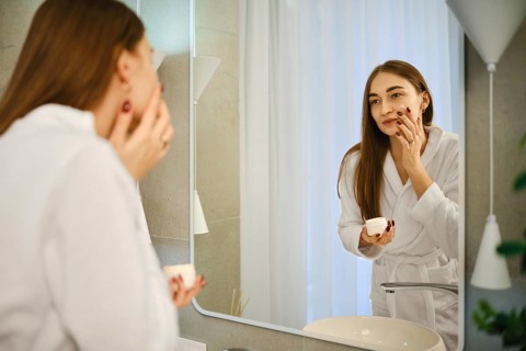 A woman stands in a bathroom wearing a robe. She is looking at her reflection in the mirror while