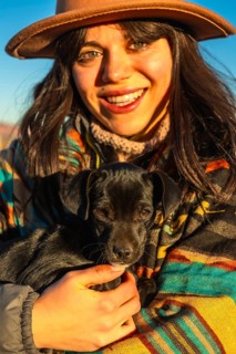Young woman wearing a hat and a colorful poncho is smiling while holding a small black puppy close,