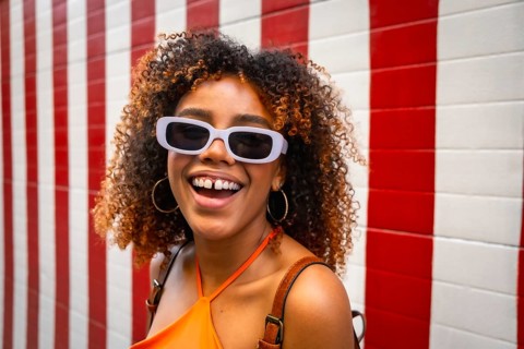 Young woman with curly brown hair wearing purple sunglasses and an orange top, laughing cheerfully