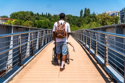 Young man with a backpack pushing a person in a wheelchair across a modern metal bridge on a sunny