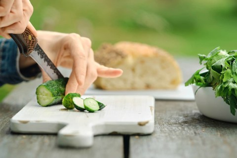 Hand holding knife cuts fresh cucumber on wooden board. Nearby, there is loaf of bread and bowl of
