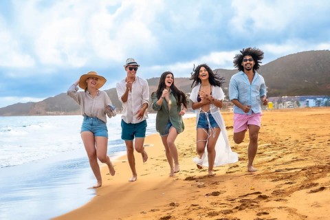 Diverse group of young adult friends running barefoot along the ocean shoreline on a sunny beach,