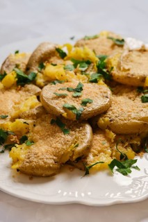 Crispy smashed crushed potatoes are arranged on a white plate. They are topped with green herbs.