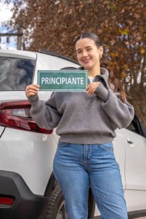 Smiling young woman holding green principiante beginner driver sign beside white SUV, outdoor