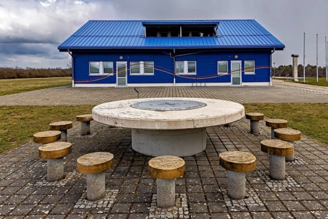 Building with blue façade and a central round table with several seats in a paved area, The Point