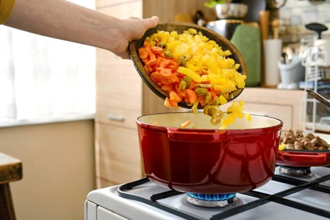 A person pours chopped vegetables into a red cooking pot placed on the stove. The kitchen setting