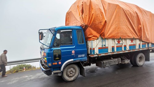 Blue truck with orange tarpaulin drives on a foggy road, isolated people visible, truck and road