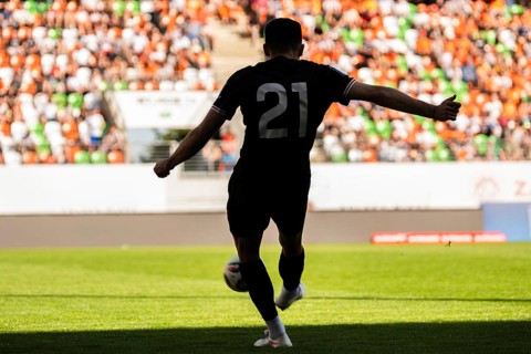Rear view of a soccer player in a black jersey kicking the ball towards the goal in a crowded