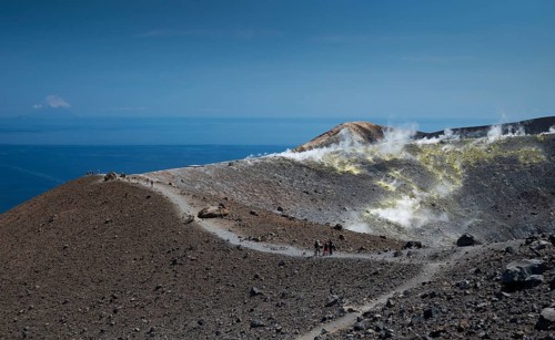 Visitors walk along the crater rim, sulfur vapors rise, in the background the Mediterranean Sea,