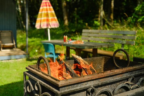 Fire pit burns wood in backyard setting. Green grass surrounds fire pit. Nearby table with drinks.