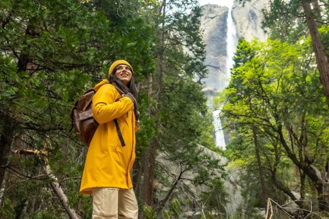 Woman in a yellow raincoat and backpack hiking through the forest, looking up with a happy