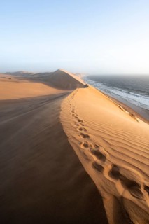 Sand dunes with wave patterns and sea in evening light, wind blowing sand across the dunes,