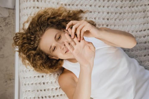 Smiling woman relaxes on bed, playing with her fingers. She has curly hair and wears a simple top.