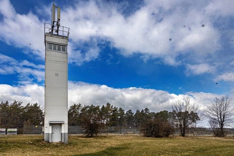 A tall watchtower rises into the blue sky, surrounded by a meadow and trees, The Point Alpha