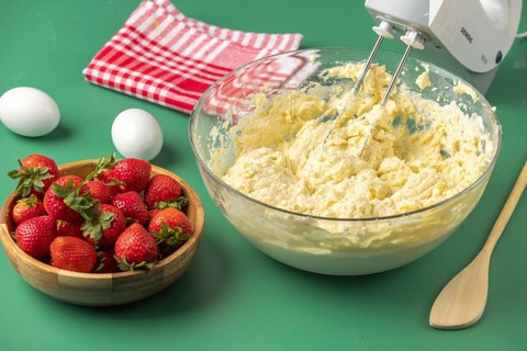A mixer whips dough in a bowl, strawberries wait in a wooden bowl