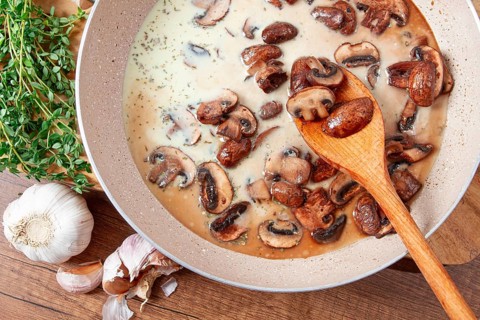 Mushrooms are cooking in a creamy sauce with garlic and herbs in a pan. Fresh thyme and garlic are