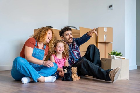 Happy family sitting on floor among moving boxes, looking away and discussing future plans together