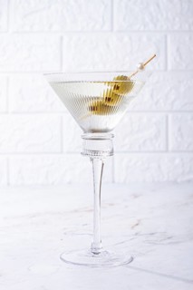 A clear glass cocktail sits on a white marble surface. Three green olives are placed on a skewer