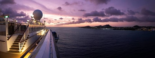 Panoramic view from a Caribbean cruise ship deck at sunset showing a purple sky over the island
