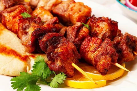 Meat skewers are placed on a white plate with slices of flatbread and lemon. Fresh herbs are