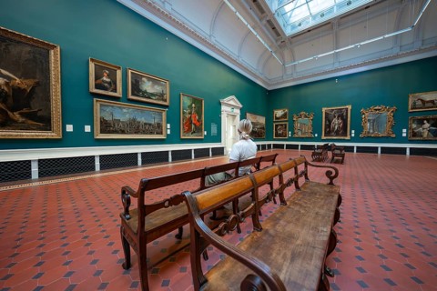 A visitor looks at paintings in an ornate gallery with green walls and wooden benches, National