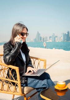 Happy business woman drinking coffee using laptop and smartphone chat. West Bay skyline background