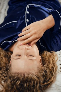 Woman with curly hair is lying on soft bed. She is wearing blue pajamas. Her hand is covering her