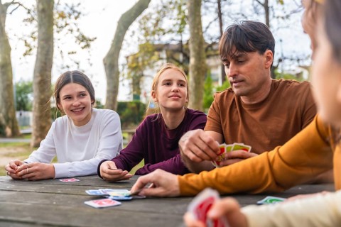Father and two daughters play cards at a park picnic table, enjoying a relaxed, sunny day of