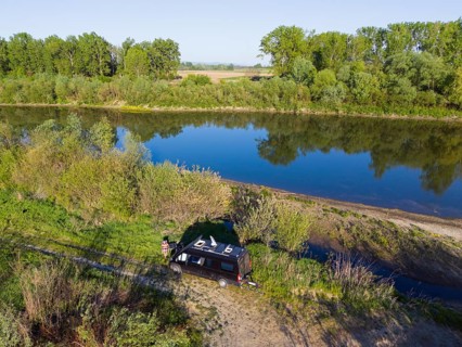 A car is parked next to a quiet river under trees, which gives a relaxed camping atmosphere, aerial