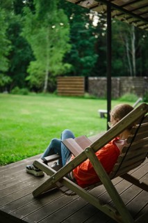 Woman sits on lounge chair in backyard surrounded by grass and trees reading book in afternoon.