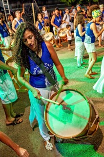 Woman in blue clothes passionately plays a big drum at a party, a band plays and dances at a