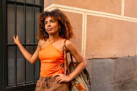 Young woman with curly hair wearing an orange top and carrying a backpack, standing next to a