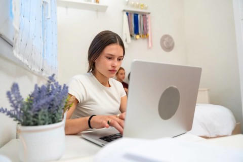 Young woman focused on her computer, working remotely or studying online from her bedroom desk,