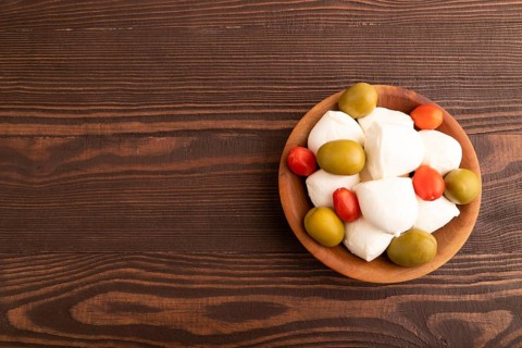 White Mozzarella cheese, with tomatoes and olives in wooden bowl on brown wooden background, top