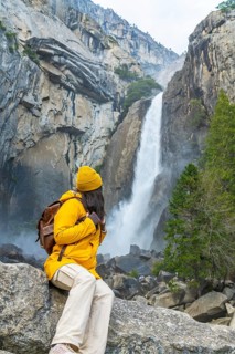 Woman in yellow jacket and beanie sits on a rock, watching lower yosemite falls plunge between