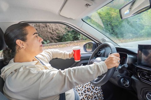 Woman driving with one hand on steering wheel while holding red coffee cup, profile view, car