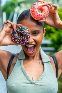 Joyful young woman with dark skin enjoying sweet donuts, holding them playfully over her eyes,